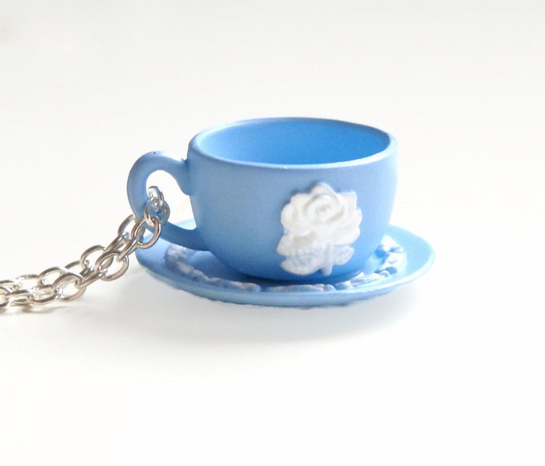 May include: A light blue teacup and saucer necklace with a white rose design. The necklace is made of a silver chain.