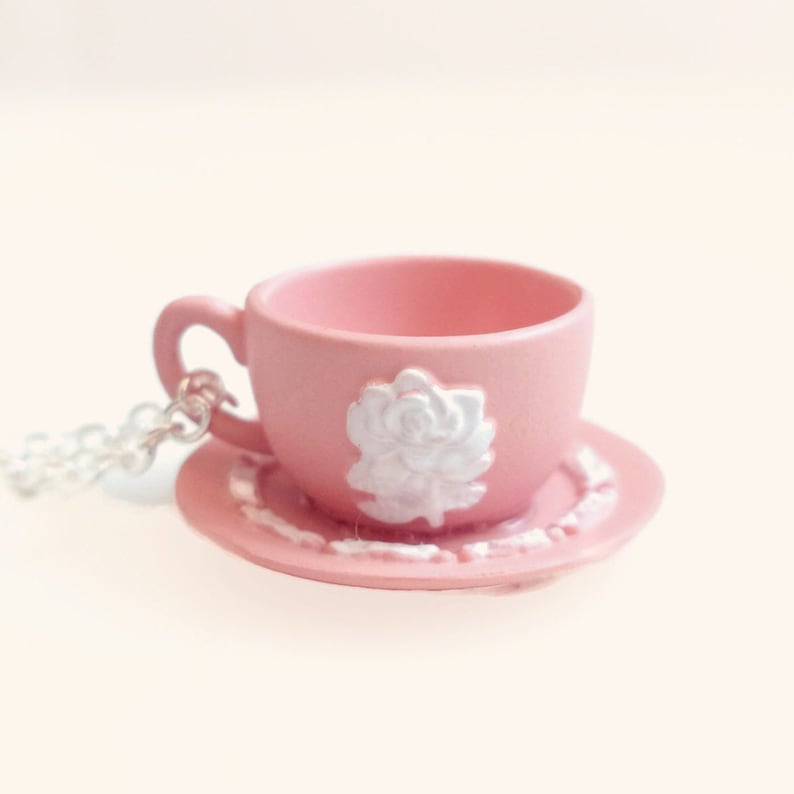 May include: A pink teacup and saucer necklace with a white rose design. The necklace is made of silver chain.