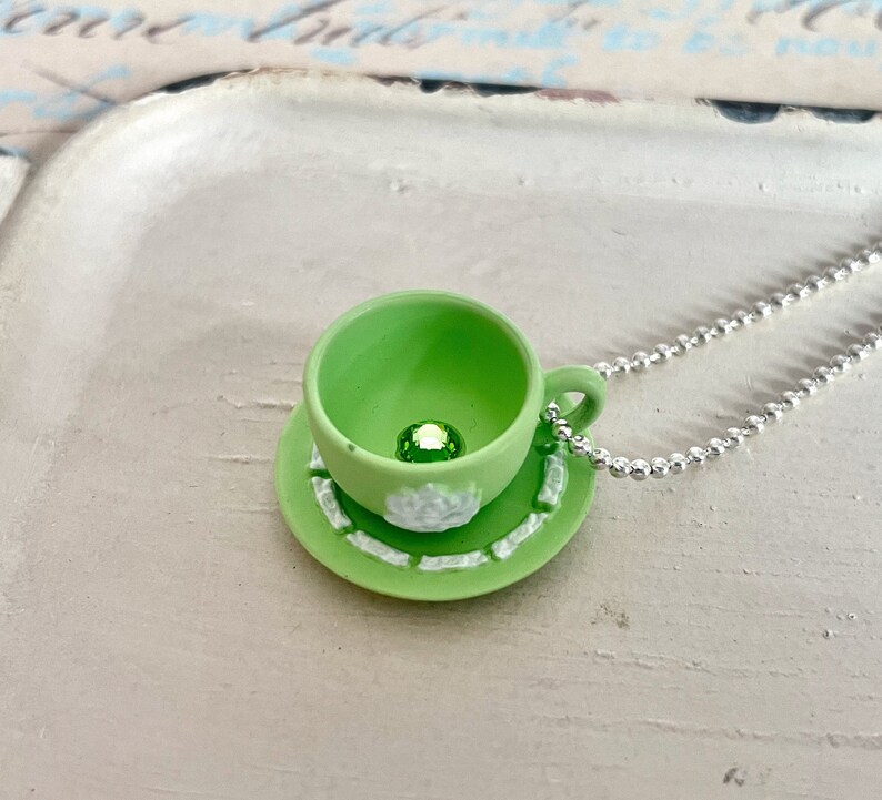 May include: A green teacup and saucer necklace with a green gem in the centre of the teacup.