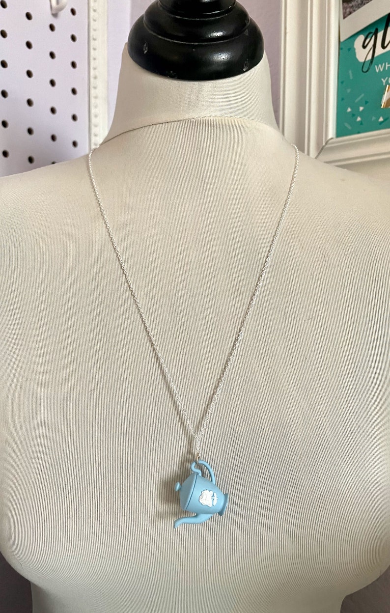 May include: A silver chain necklace with a light blue teapot pendant. The teapot has a white floral design.