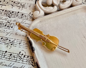 May include: A gold-colored violin brooch on a white surface. The brooch is detailed with a string design and a small bow.