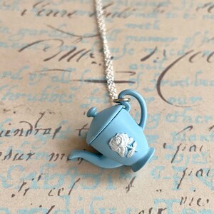 May include: A light blue teapot charm necklace with a white rose design. The charm is attached to a silver chain.