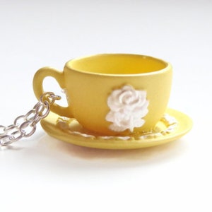 May include: A yellow teacup and saucer necklace with a white rose design. The chain is silver.