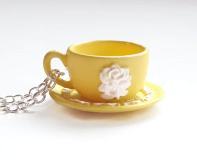May include: A yellow teacup and saucer necklace with a white rose design. The chain is silver.