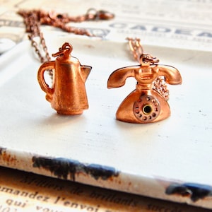 May include: A copper necklace with two charms: a vintage-style rotary phone and a coffee pot. The charms are detailed and have a warm, aged look.