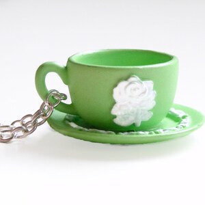 May include: A green teacup and saucer necklace with a white rose design. The necklace is made of a silver chain.