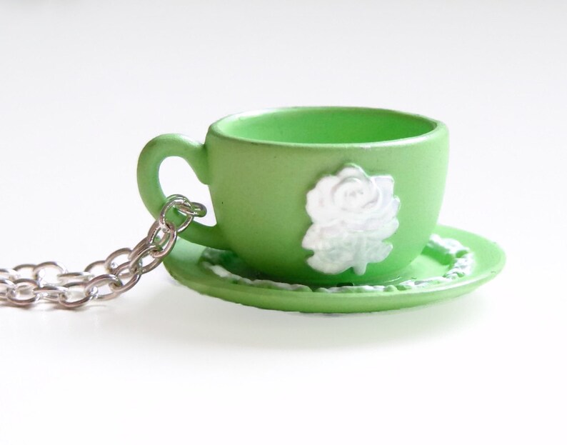 May include: A green teacup and saucer necklace with a white rose design. The necklace is made of a silver chain.