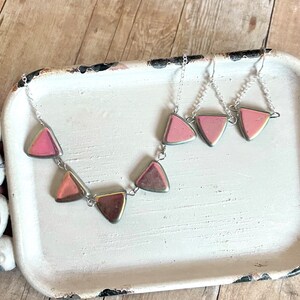 Puede incluir: Un collar y pendientes de cadena plateada con colgantes de piedra preciosa triangular rosa. El collar tiene cinco colgantes y los pendientes tienen tres colgantes.