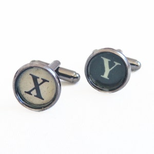 Può includere: Un paio di gemelli con tasti di macchina da scrivere vintage. Il gemello sinistro ha la lettera 'X' su un tasto color crema, e il gemello destro ha la lettera 'Y' su un tasto verde scuro.