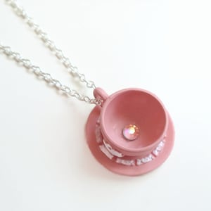 May include: A pink teacup and saucer necklace with a clear rhinestone in the centre of the teacup. The necklace is made of a silver chain.