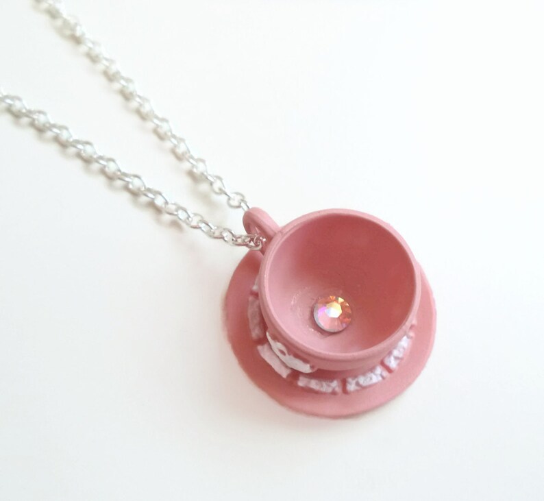 May include: A pink teacup and saucer necklace with a clear rhinestone in the centre of the teacup. The necklace is made of a silver chain.