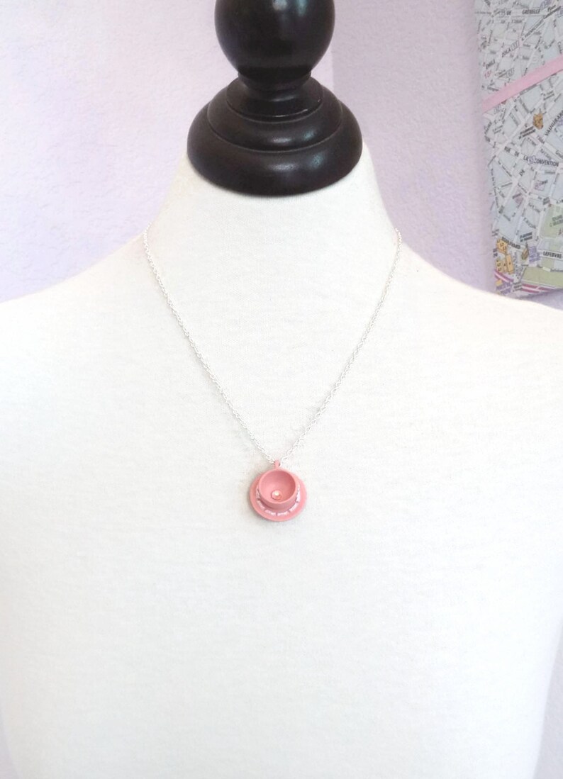 May include: A delicate silver chain necklace with a pink teacup pendant. The teacup has a white rim and a small pearl in the centre.