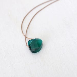 May include: A heart-shaped, emerald-green gemstone pendant necklace. The pendant is faceted and suspended from a fine, gold-toned chain. The necklace is presented on a white surface.