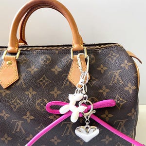 May include: A brown handbag with a repeating pattern, tan leather handles, and gold-tone hardware. A pink strap and silver charm with a white heart and a white dog-shaped charm hang from the handle.