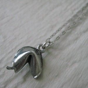 May include: A silver-colored fortune cookie pendant necklace. The pendant is a detailed replica of a fortune cookie, with a slightly open design. The necklace chain is delicate and silver, complementing the pendant's color.