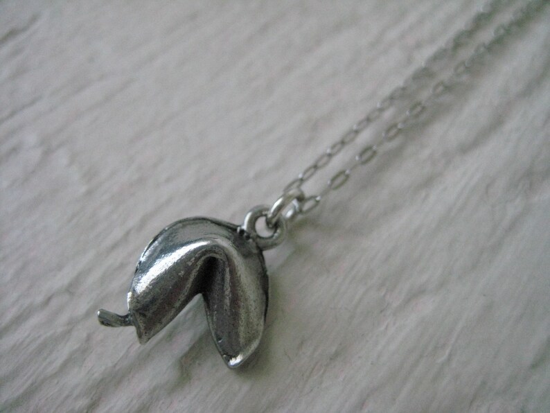 May include: A silver-colored fortune cookie pendant necklace. The pendant is a detailed replica of a fortune cookie, with a slightly open design. The necklace chain is delicate and silver, complementing the pendant's color.