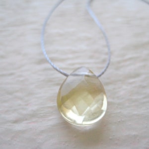 Puede incluir: Un colgante transparente en forma de l&aacute;grima con un tono amarillo claro que cuelga de un cord&oacute;n blanco.