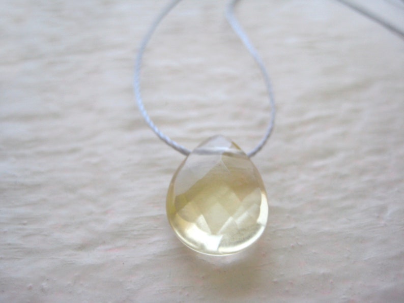 Puede incluir: Un colgante transparente en forma de l&aacute;grima con un tono amarillo claro que cuelga de un cord&oacute;n blanco.