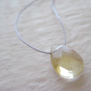 Puede incluir: Un collar con un colgante en forma de l&aacute;grima, hecho de una piedra preciosa transparente de color amarillo claro. El colgante est&aacute; suspendido de un cord&oacute;n blanco delgado.