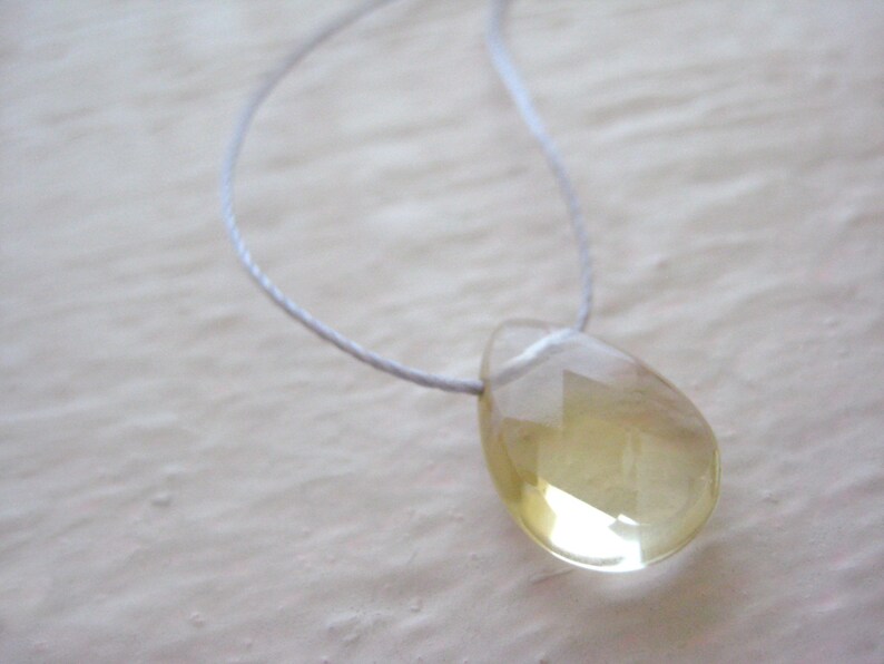 Puede incluir: Un collar con un colgante en forma de l&aacute;grima, hecho de una piedra preciosa transparente de color amarillo claro. El colgante est&aacute; suspendido de un cord&oacute;n blanco delgado.