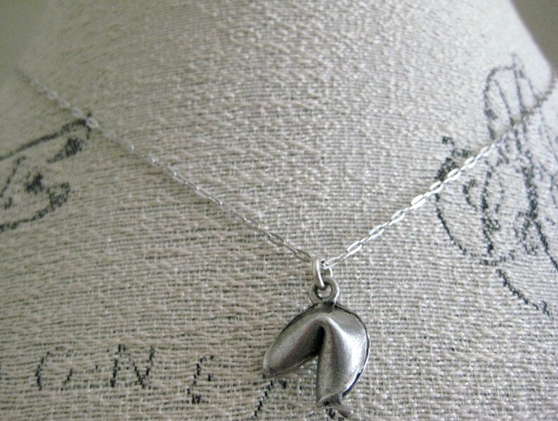 May include: A silver-colored necklace featuring a fortune cookie pendant. The pendant hangs from a delicate chain. The necklace is displayed against a textured, off-white background.