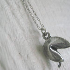 May include: A silver-colored fortune cookie pendant hangs from a delicate chain. The pendant is detailed with a textured surface, and the cookie is slightly open. The necklace is displayed against a white, textured background.