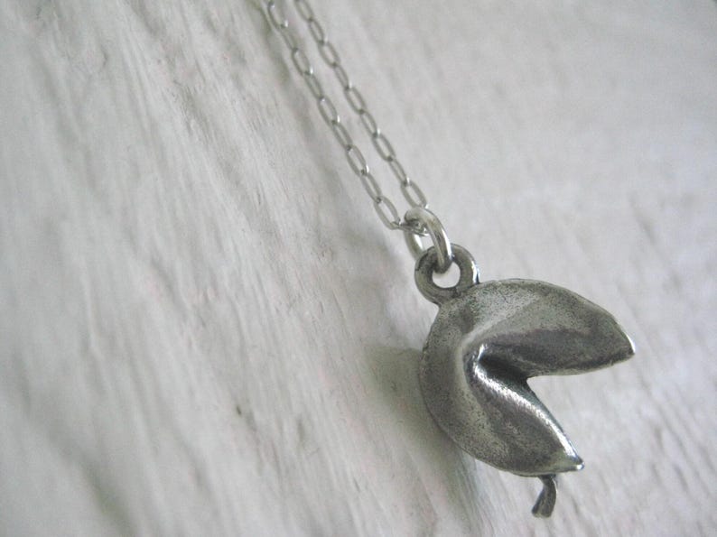 May include: A silver-colored fortune cookie pendant hangs from a delicate chain. The pendant is detailed with a textured surface, and the cookie is slightly open. The necklace is displayed against a white, textured background.