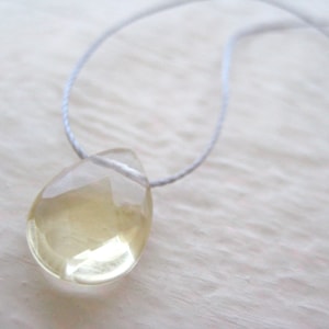 Puede incluir: Un collar con un colgante en forma de l&aacute;grima, hecho de una piedra preciosa transparente de color amarillo claro. El colgante est&aacute; suspendido de un cord&oacute;n fino y blanco.