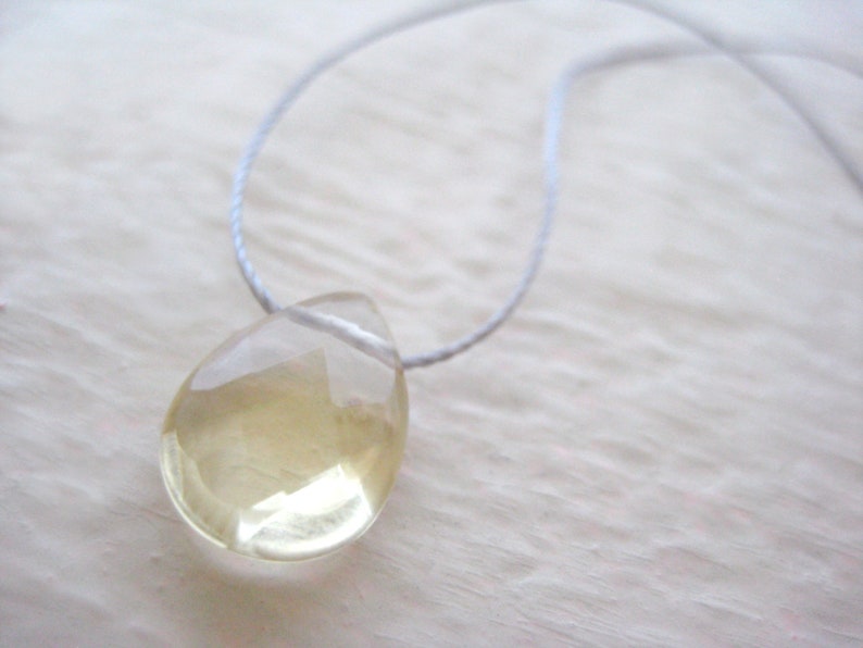 Puede incluir: Un collar con un colgante en forma de l&aacute;grima, hecho de una piedra preciosa transparente de color amarillo claro. El colgante est&aacute; suspendido de un cord&oacute;n fino y blanco.