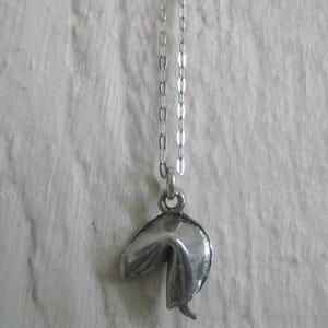 May include: A silver-colored fortune cookie pendant hangs from a delicate chain. The pendant is detailed with realistic folds and creases, giving it a textured appearance. The necklace is displayed against a textured white background.