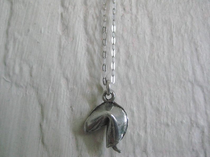 May include: A silver-colored fortune cookie pendant hangs from a delicate chain. The pendant is detailed with realistic folds and creases, giving it a textured appearance. The necklace is displayed against a textured white background.