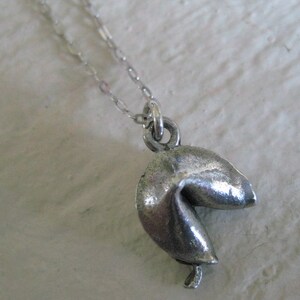May include: A silver-colored fortune cookie pendant hangs from a delicate chain. The pendant is detailed with realistic folds and creases, giving it a textured appearance. The chain is a fine, link style, and the pendant is suspended from a small loop.