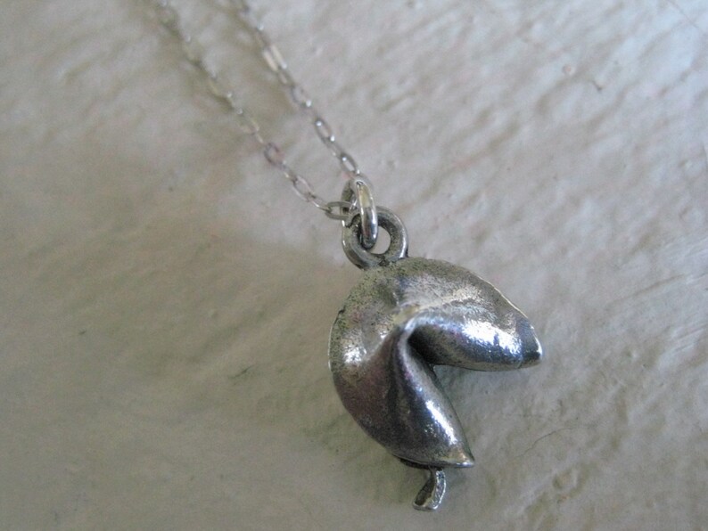 May include: A silver-colored fortune cookie pendant hangs from a delicate chain. The pendant is detailed with realistic folds and creases, giving it a textured appearance. The chain is a fine, link style, and the pendant is suspended from a small loop.