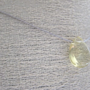 Puede incluir: Un colgante transparente en forma de l&aacute;grima cuelga de un cord&oacute;n blanco fino. El colgante est&aacute; hecho de un material amarillo transl&uacute;cido, posiblemente citrino o topacio.