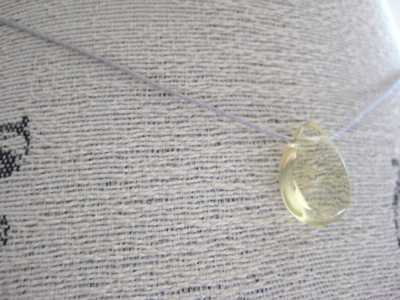 Puede incluir: Un colgante transparente en forma de l&aacute;grima cuelga de un cord&oacute;n blanco fino. El colgante est&aacute; hecho de un material amarillo transl&uacute;cido, posiblemente citrino o topacio.