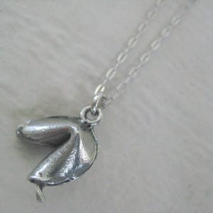 May include: A silver-colored fortune cookie pendant necklace. The pendant is detailed with textured lines and hangs from a delicate chain. The necklace is displayed against a soft, white background.