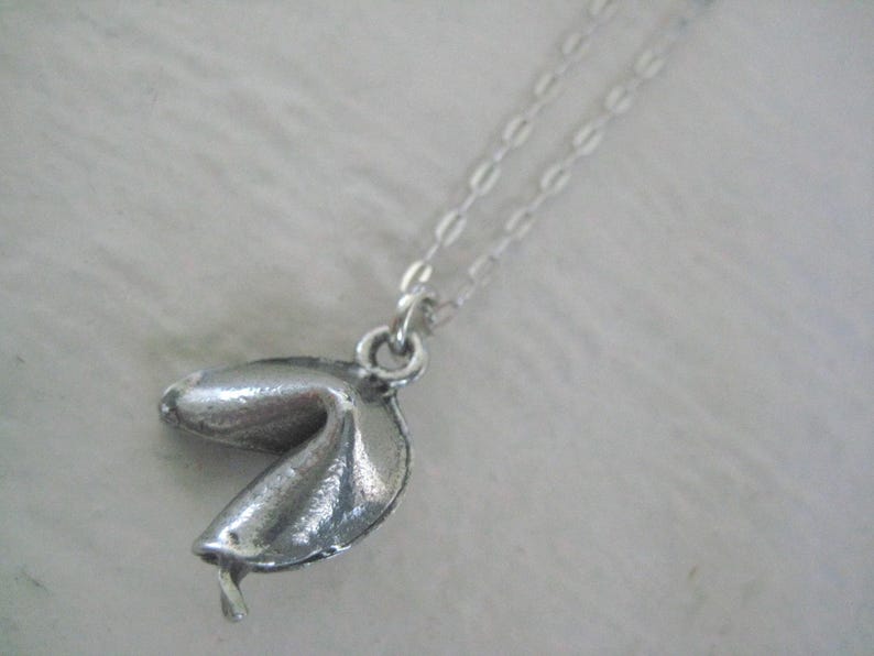 May include: A silver-colored fortune cookie pendant necklace. The pendant is detailed with textured lines and hangs from a delicate chain. The necklace is displayed against a soft, white background.