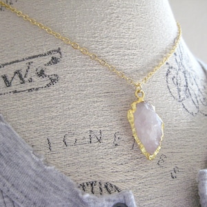 May include: A gold-colored chain necklace with a pendant featuring a light pink crystal with gold-colored edging. The necklace is displayed on a mannequin wearing a gray shirt.