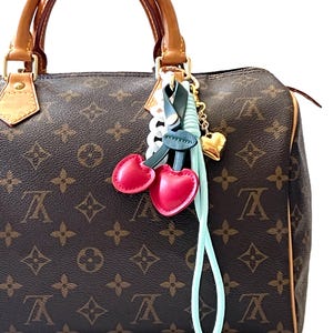 May include: A brown monogrammed handbag with tan leather handles and trim, featuring a decorative charm attached to the handle. The charm includes two red heart-shaped pieces, a dark green strap, a white chain link, and a gold-toned heart pendant.