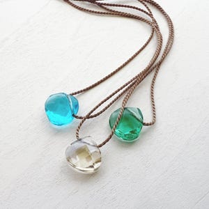 May include: Three necklaces featuring faceted, heart-shaped pendants in translucent blue, green, and clear glass. Each pendant is suspended from a thin, brown twisted cord. The pendants have a polished, reflective surface that catches light, creating a clear, gem-like appearance against the cord material.