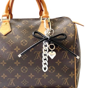 May include: A brown monogram patterned handbag with tan leather handles and trim. A decorative silver-toned metal keychain hangs from the handle, featuring a black leather bow, a thick silver chain link, and a small silver heart-shaped charm.