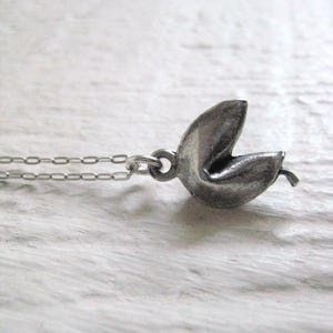 May include: A silver-colored fortune cookie pendant on a delicate chain. The pendant is detailed with a realistic texture, and the chain is a simple link style. The necklace is displayed against a light-colored, textured background.