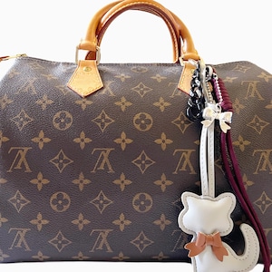 May include: A brown monogram print handbag with tan leather handles and a zipper closure. A decorative charm hangs from the handle, featuring a white cat-shaped leather piece, a silver bow, a black chain link, and a dark red braided cord.