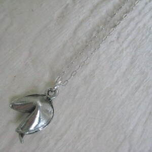 May include: Silver-toned necklace featuring a fortune cookie pendant. The pendant is detailed with textured surfaces and a realistic shape. The chain is delicate and complements the pendant's design. A unique accessory for everyday wear.