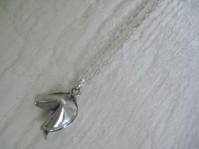 May include: Silver-toned necklace featuring a fortune cookie pendant. The pendant is detailed with textured surfaces and a realistic shape. The chain is delicate and complements the pendant's design. A unique accessory for everyday wear.