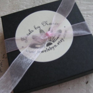 Pu&ograve; includere: Una scatola regalo nera con un nastro rosa e un'etichetta bianca che dice "Jewels by Nancy" e "welbynn.etsy.com".