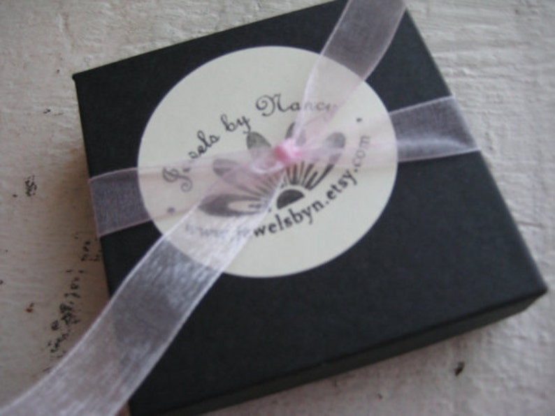 Pu&ograve; includere: Una scatola regalo nera con un nastro rosa e un'etichetta bianca che dice "Jewels by Nancy" e "welbynn.etsy.com".