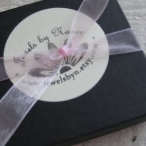 Puede incluir: Una caja de regalo negra con una etiqueta circular blanca que dice "Jewels by Nancy" y "welsbyn.etsy.com". La caja est&aacute; atada con una cinta rosa.