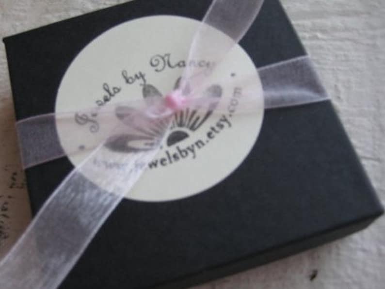 Puede incluir: Una caja de regalo negra con una etiqueta circular blanca que dice "Jewels by Nancy" y "welsbyn.etsy.com". La caja est&aacute; atada con una cinta rosa.