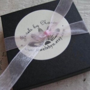 May include: A black square gift box with a white circular label and a sheer ribbon. The label reads "Jewels by Nancy" and "welsbyn.etsy.com". The ribbon is wrapped around the box and tied in a bow.